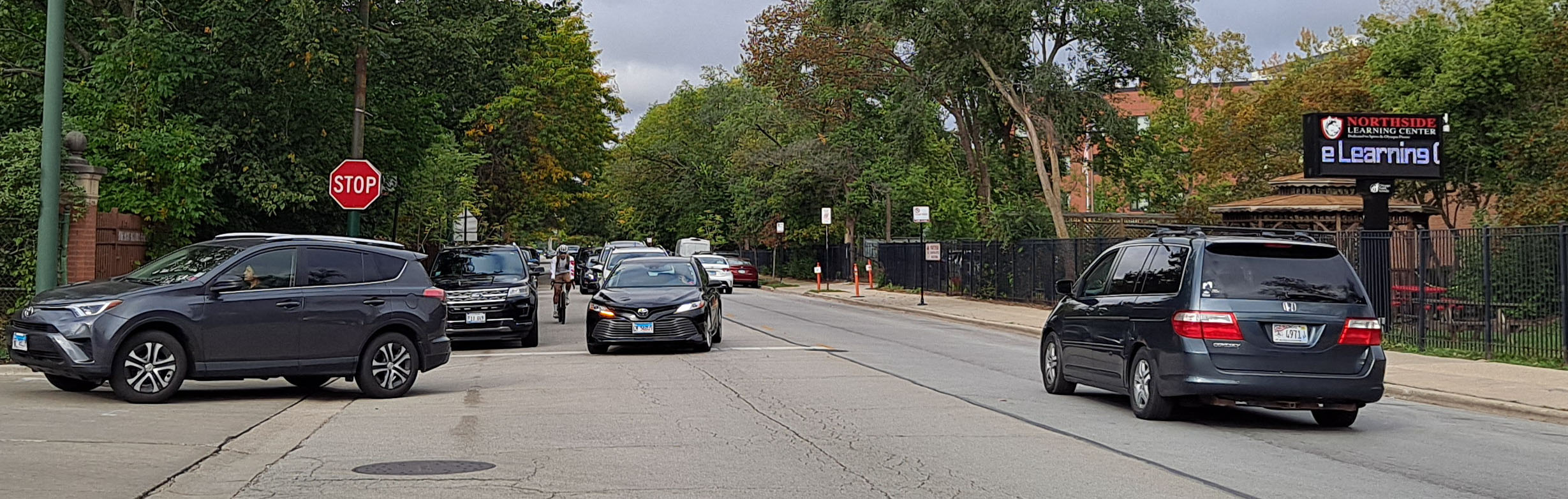 View of intersection with family posed to cross street, car traveling parallel to family, and cyclist riding on roadway.
