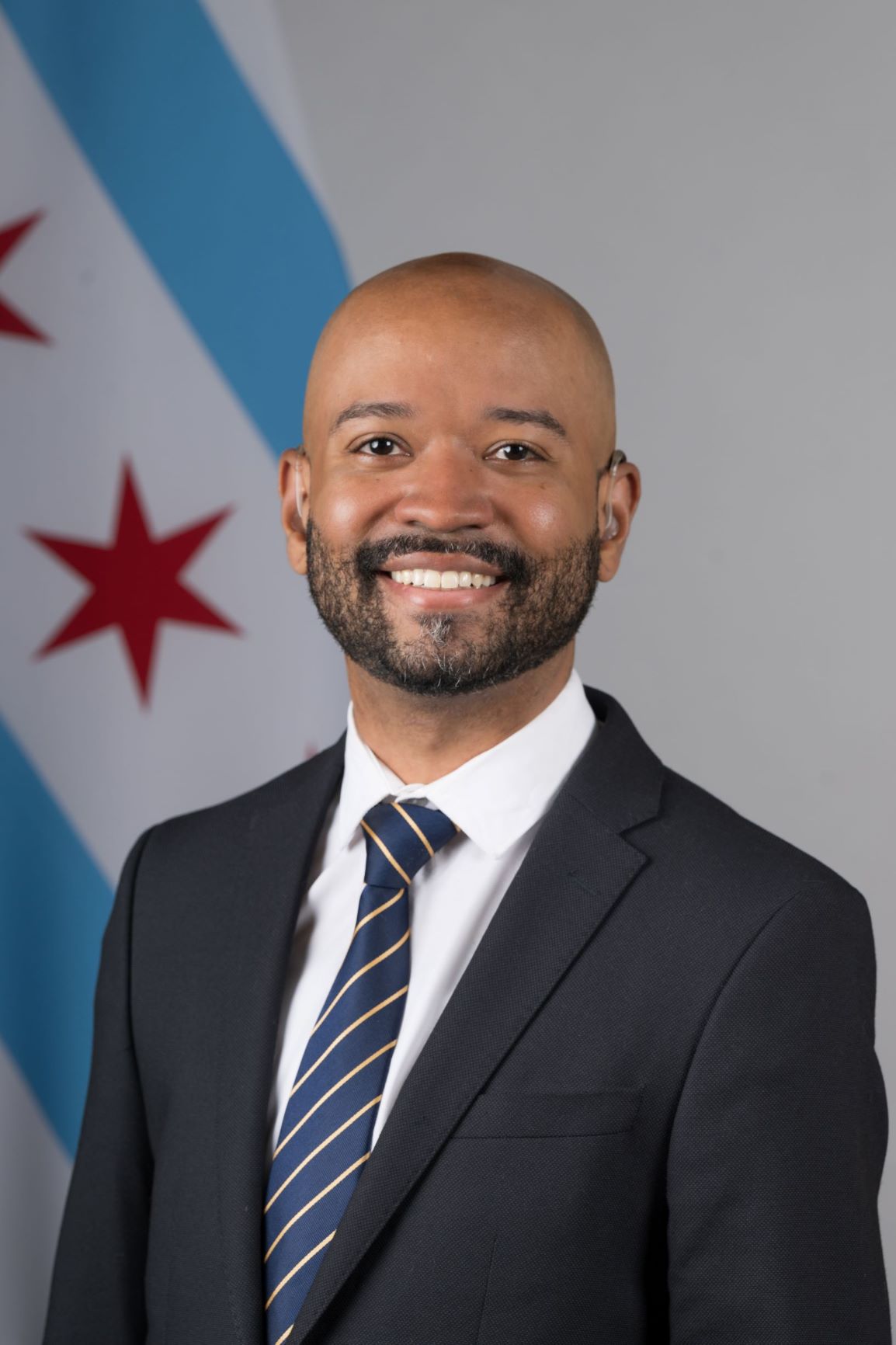 Project Coordinator Nathaniel Kelly is seen in his City headshot with a suit in front of the City of Chicago flag.