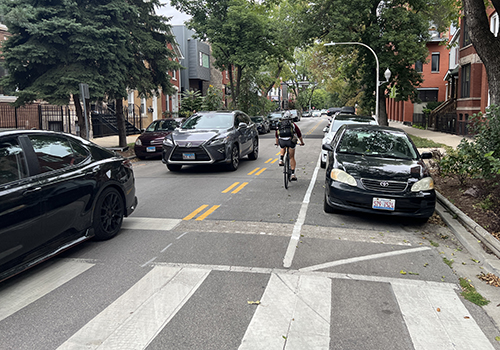 Person biking towards viewer with in a bike lane with a double dashed yellow line separating the cyclist from oncoming vehicle traffic.