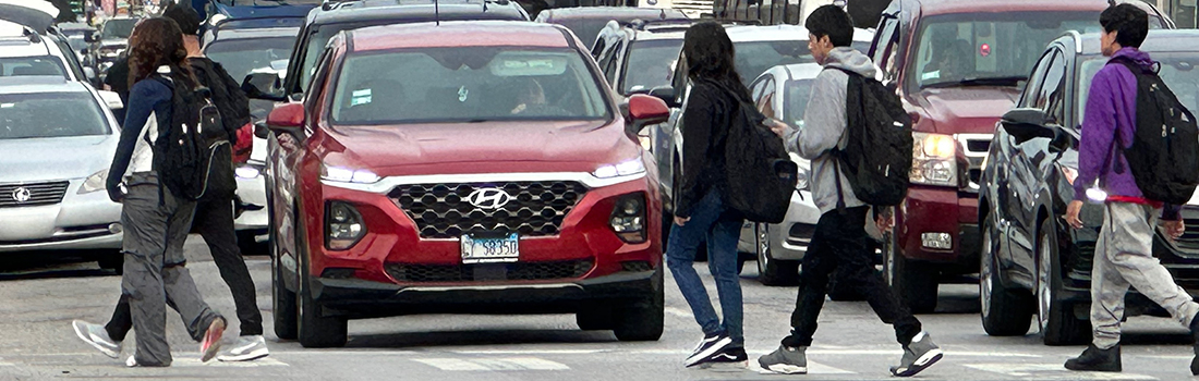 View of intersection with family posed to cross street, car traveling parallel to family, and cyclist riding on roadway.