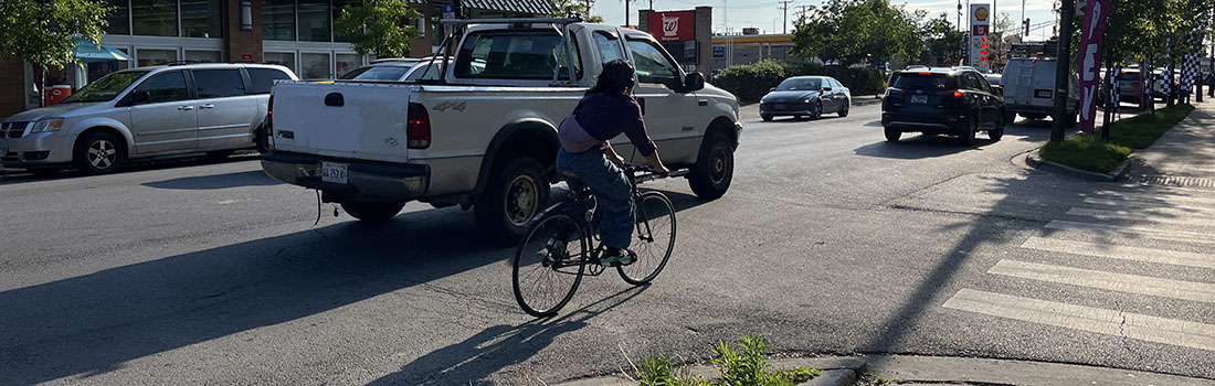 View of intersection with family posed to cross street, car traveling parallel to family, and cyclist riding on roadway.