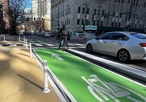 Cyclist stopped at intersection red light facing away from camera in green-painted bike lane with concrete separating it from motor vehicle.