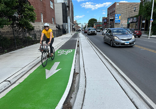 Person biking towards viewer in green painted bike lane separated from vehicle travel lane by concrete curb.