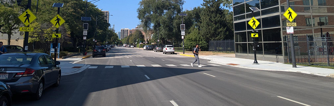 View of intersection with family posed to cross street, car traveling parallel to family, and cyclist riding on roadway.