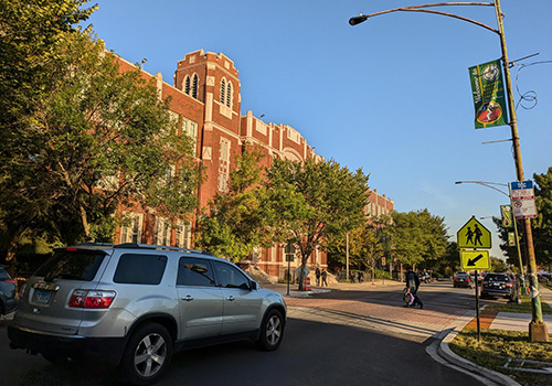 Image of street in front of kelly high school with a motorist yielding to a pedestrian crossing the street in a raised crosswalk