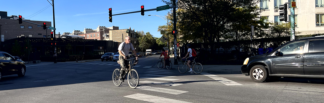 View of intersection with family posed to cross street, car traveling parallel to family, and cyclist riding on roadway.