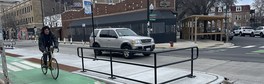 View of intersection with family posed to cross street, car traveling parallel to family, and cyclist riding on roadway.