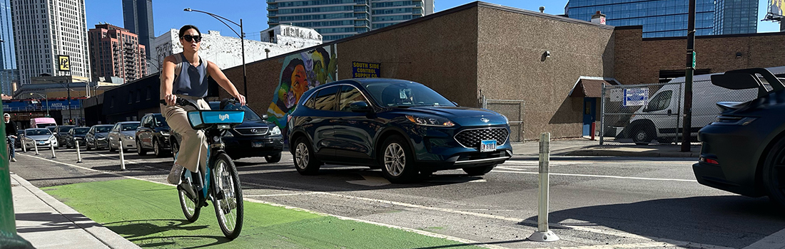 View of intersection with family posed to cross street, car traveling parallel to family, and cyclist riding on roadway.