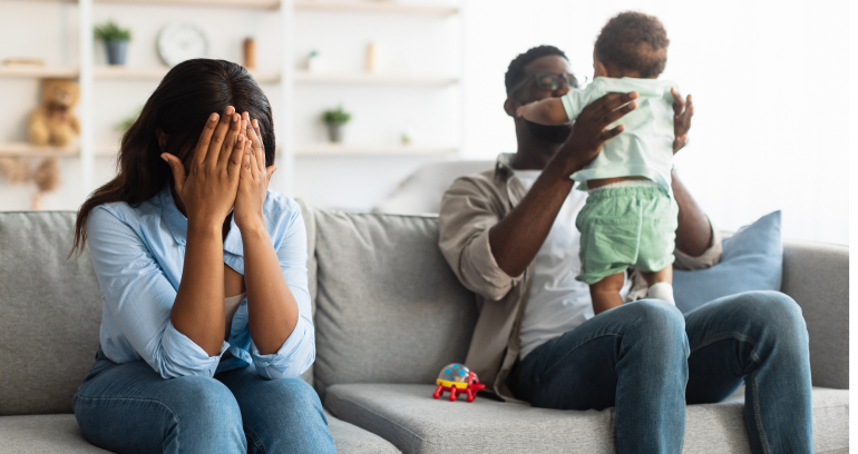 Mother seated on couch with hands over face, father playing with baby at right